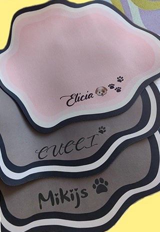 Three personalised feeding mats stacked, showing pink, grey and dark grey